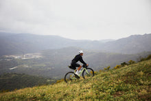 Load image into Gallery viewer, The Vittoria Terreno T30 gravel tyre,  Tubeless Ready  Person riding a bicycle on a grassy hill with a mountainous landscape in the background